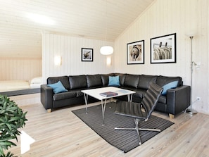 Een moderne woonkamer met een zwarte leren modulaire bank, een witte salontafel en een zwart-grijze fauteuil. Op de muur hangen diverse ingekaderde foto’s, er staat een vloerlamp en een potplant.