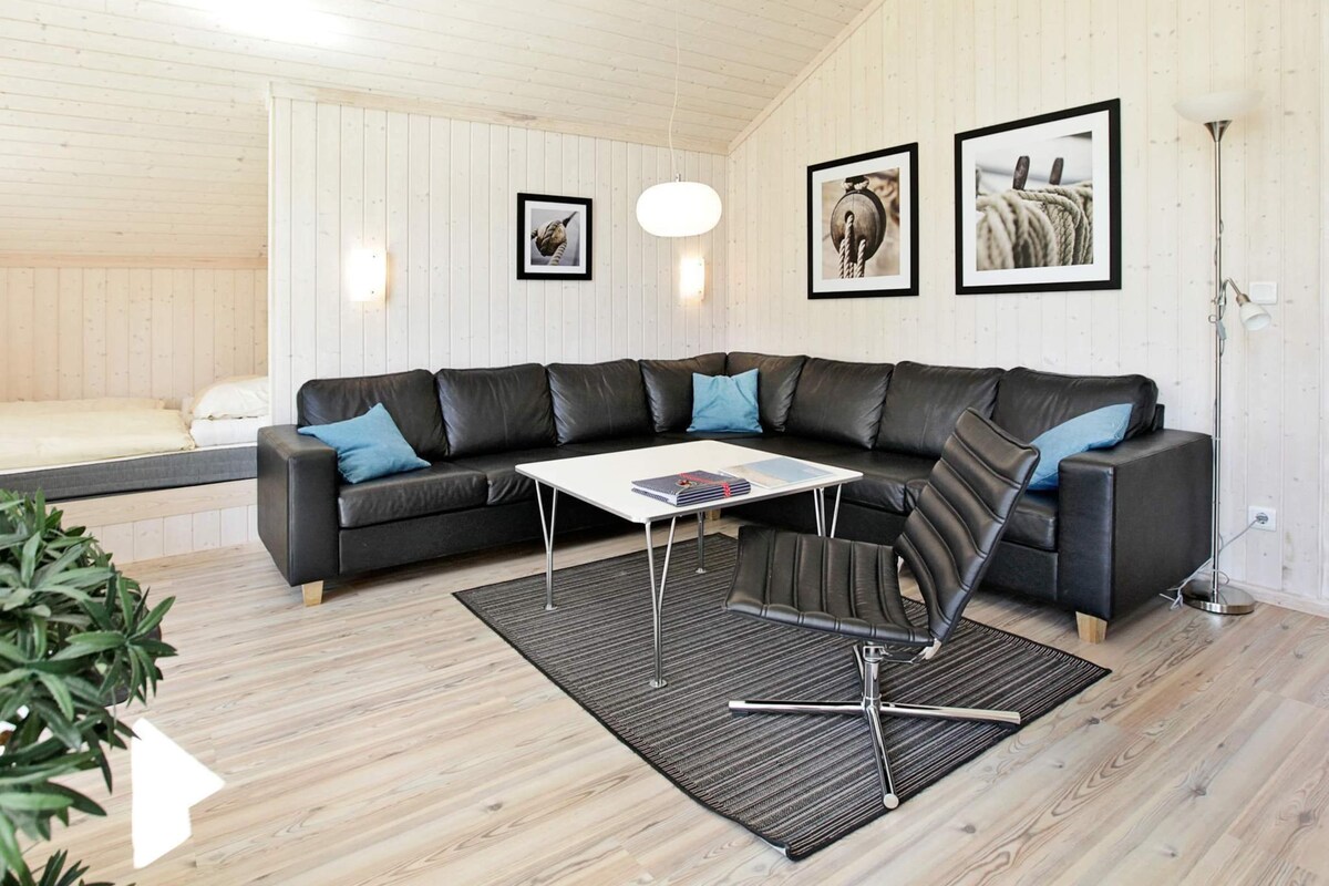 Ein modernes Wohnzimmer mit einem schwarzen Ledersofa, einem weißen Couchtisch und einem schwarz-weißen Teppich. An der Wand hängen drei gerahmte Bilder und es steht eine Stehlampe.