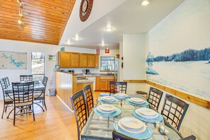 Un coin repas avec une table en verre dressée pour six personnes, entourée de chaises en bois, dans une pièce dotée de plafonds en bois et d’un grand tableau représentant un paysage enneigé au mur.