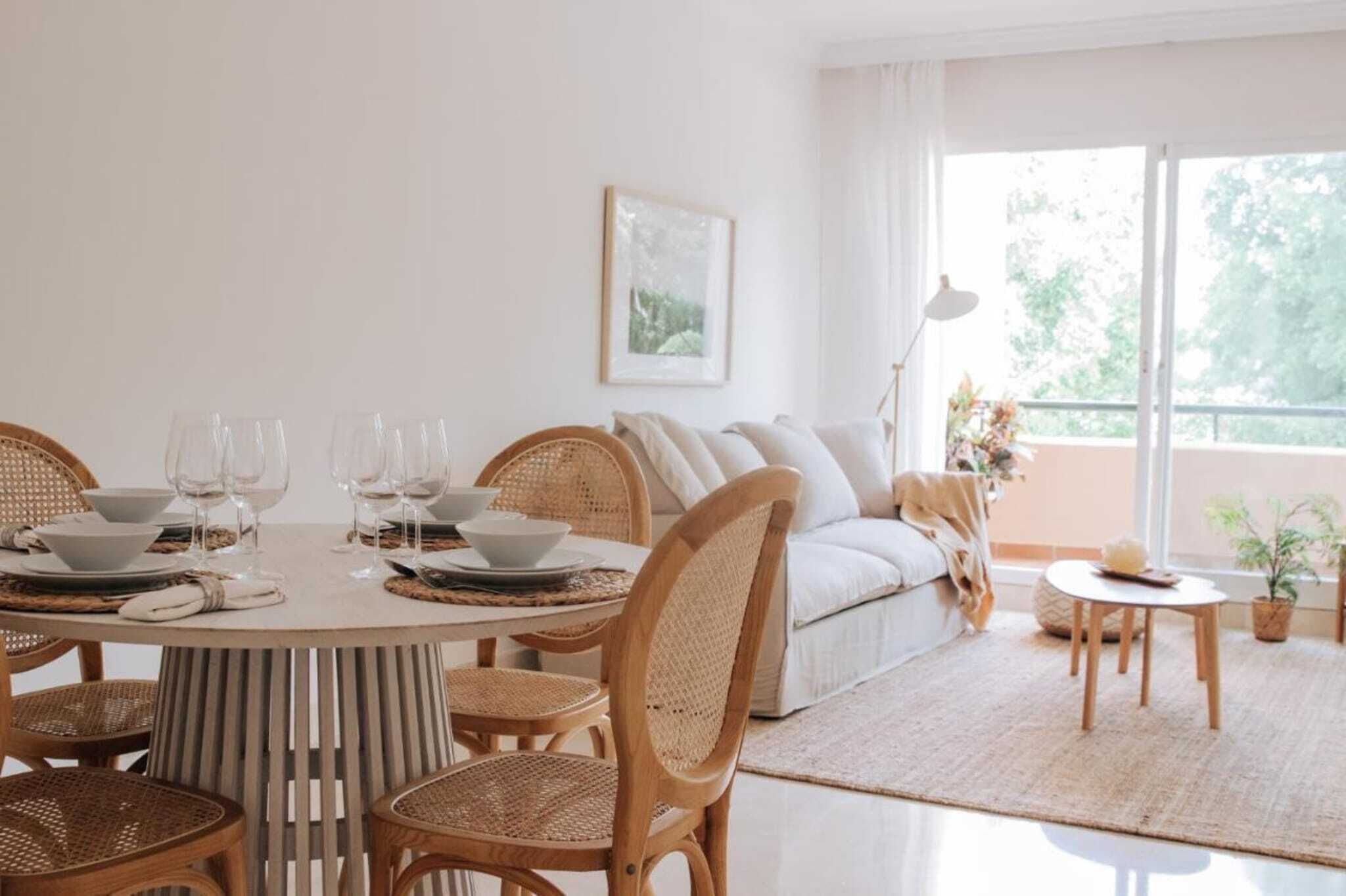 Ein Essbereich mit einem runden Tisch, gedeckt für vier Personen, umgeben von Korbstühlen. Im Hintergrund sind eine Couch und ein kleiner runder Tisch mit einer Lampe zu sehen.
