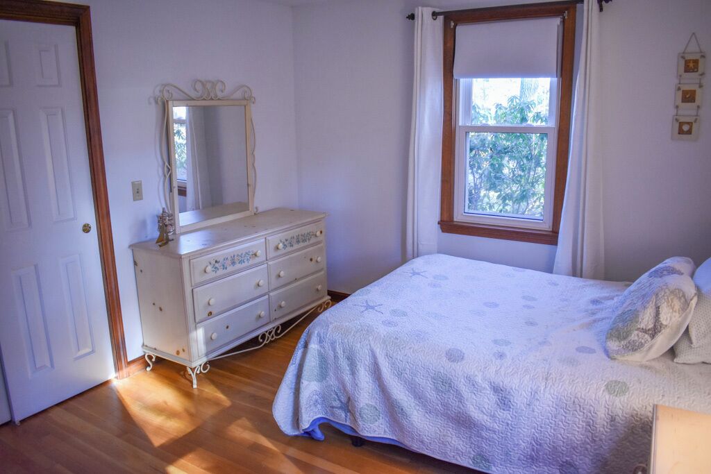 Ein Schlafzimmer mit einem Holzkoffer, einem Bett mit gemustertem Überwurf und einem Fenster mit Blick auf Bäume.