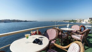 A deck with wicker chairs and a table, overlooking a body of water with buildings in the distance. - Orient Nile Romance Nile Cruise - Every Saturday from Luxor for 07 & 04 Nights - Every Wednesday From Aswan for 03 Nights (Luxor)