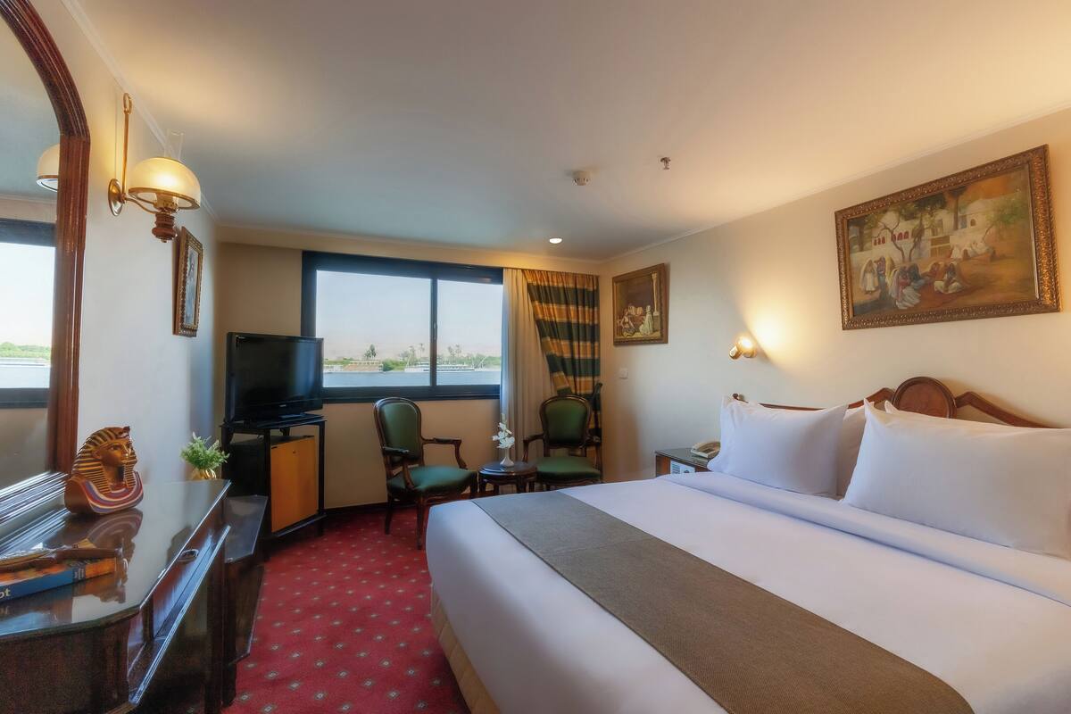 A hotel room with a large bed, a desk with a television, two chairs, a small table with a plant, and a painting on the wall.