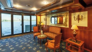 A room with a sofa, chairs, a table, and a lamp. There is a mirror on the wall and a door leading to a balcony. - Orient Nile Romance Nile Cruise - Every Saturday from Luxor for 07 & 04 Nights - Every Wednesday From Aswan for 03 Nights (Luxor)