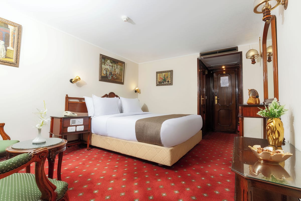 A hotel room with a bed, a desk, a chair, a small table with a vase, and a mirror.