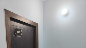 Una puerta de madera oscura con una placa numerada, dos plantas en macetas y una lámpara en la pared.