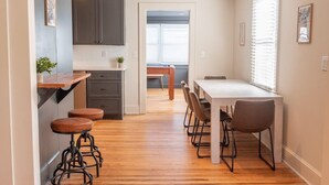 A modern kitchen with wooden flooring, dark cabinets, a dining table with chairs, and a small bar area with stools. - A Walk in the Pines - Bright & Cute 3br 2ba Home (North Charleston)