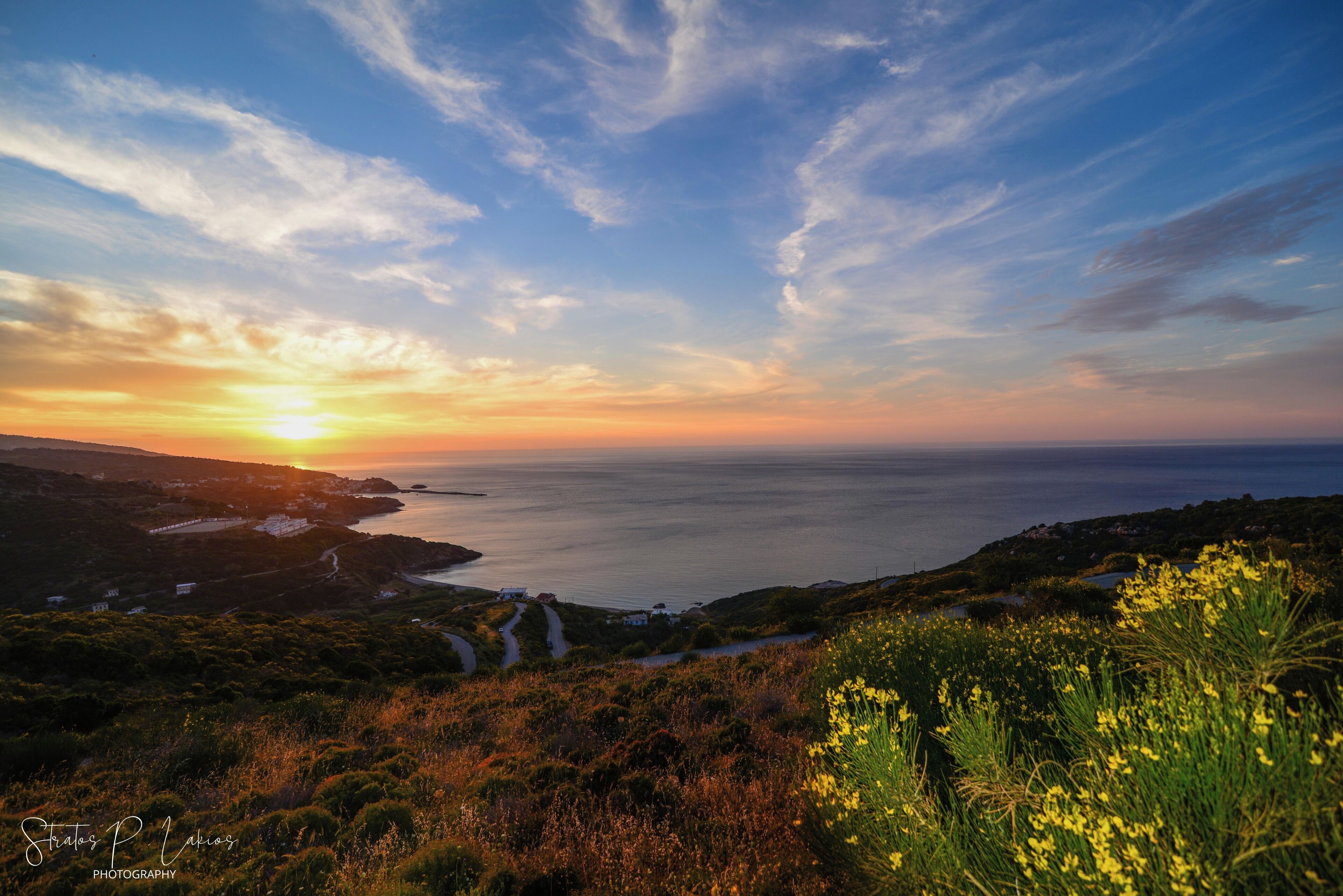 A coastal landscape with a winding road, greenery, and a sunset over the sea.