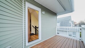 Een houten terras met een witte balustrade, een deur die leidt naar een binnenruimte met een trap, en een lichtgroene buitenmuur.