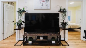 Een woonkamer met een grote flatscreen televisie, houten vloeren en potplanten op standaardjes.