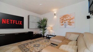 Uma sala de estar com uma televisão de tela plana grande exibindo 'NETFLIX', um sofá, um tapete estampado, um vaso de planta e uma luminária fixada na parede.