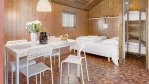 Una habitación acogedora con paredes de madera y suelo de baldosas, que incluye una mesa de comedor con sillas, una cama con sábanas blancas y una lámpara colgante.