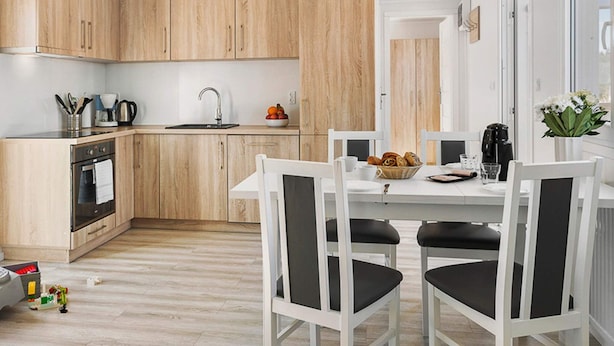 Una cocina moderna con armarios de madera, horno integrado y comedor con una mesa blanca y sillas negras.
