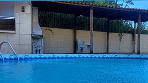 Une piscine avec un bord carrelé, un escalier en métal et un espace abrité équipé de chaises.