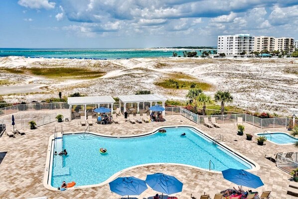 A pool area with lounge chairs and umbrellas, surrounded by a sandy landscape and a view of the ocean.
