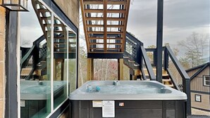 A hot tub on a wooden deck with a wooden ladder and railing, surrounded by a building with large windows.