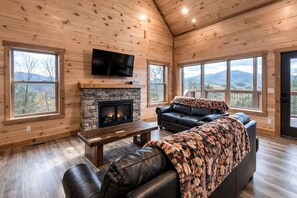 A cozy cabin interior with a stone fireplace, leather seating, and large windows offering mountain views. - Breathless Views | Private Heated Pool, Hot Tub, Pet Friendly! (Sevierville)