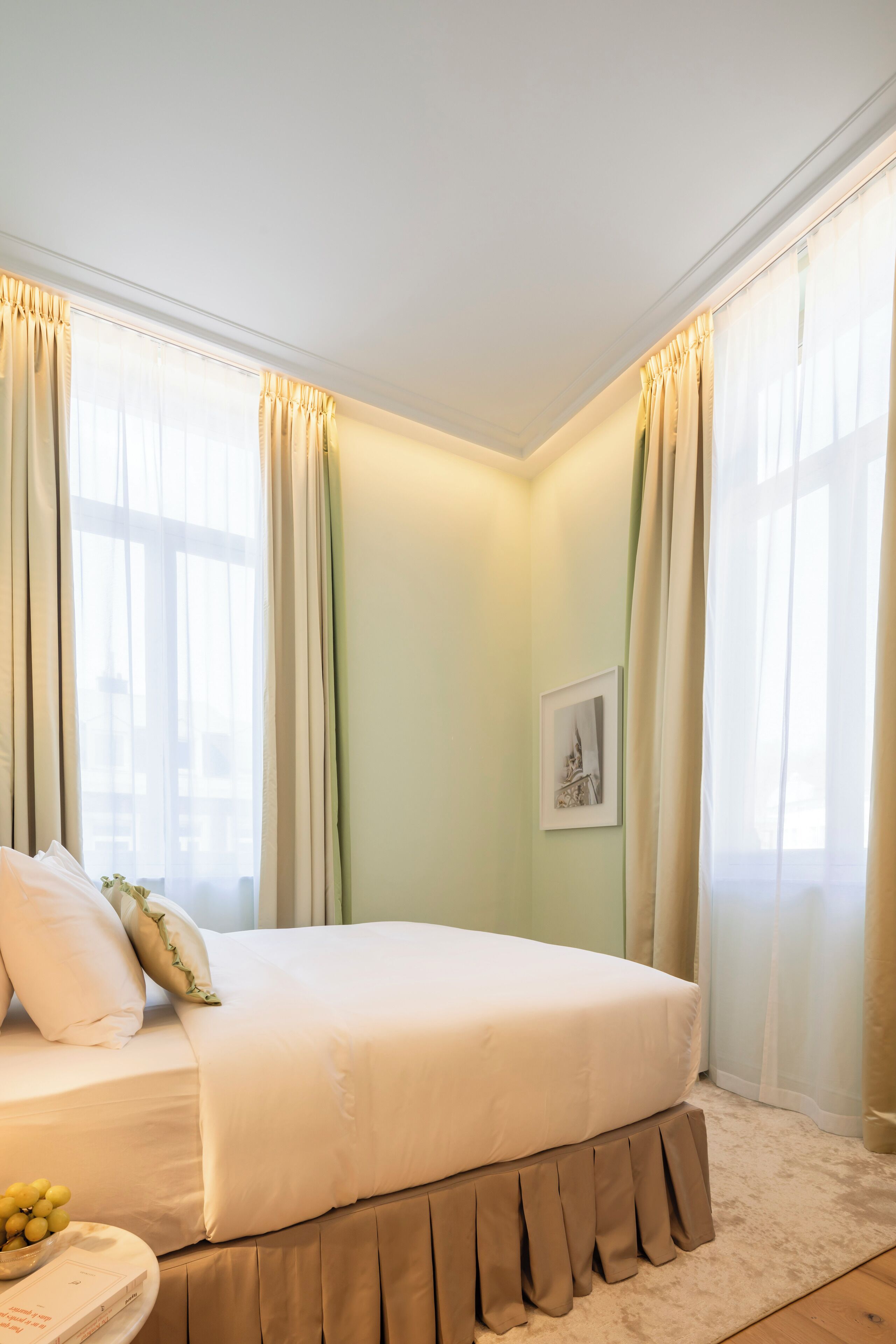 A neatly arranged bedroom with a large bed, a bedside table with a book and a bowl of fruit, and a framed picture on the wall.
