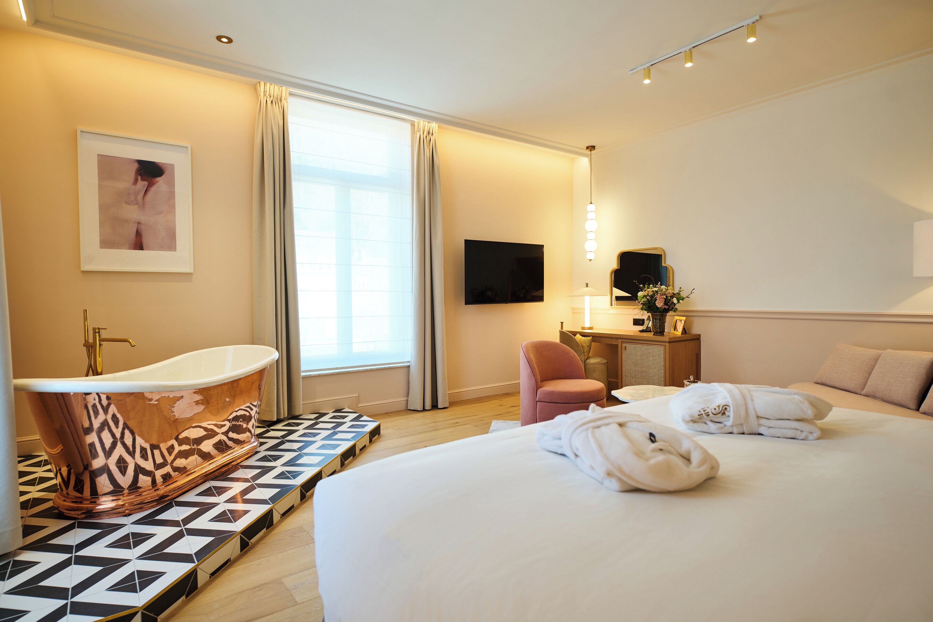 A modern hotel room with a large bed, a bathtub, a desk with a lamp, and a TV on the wall.