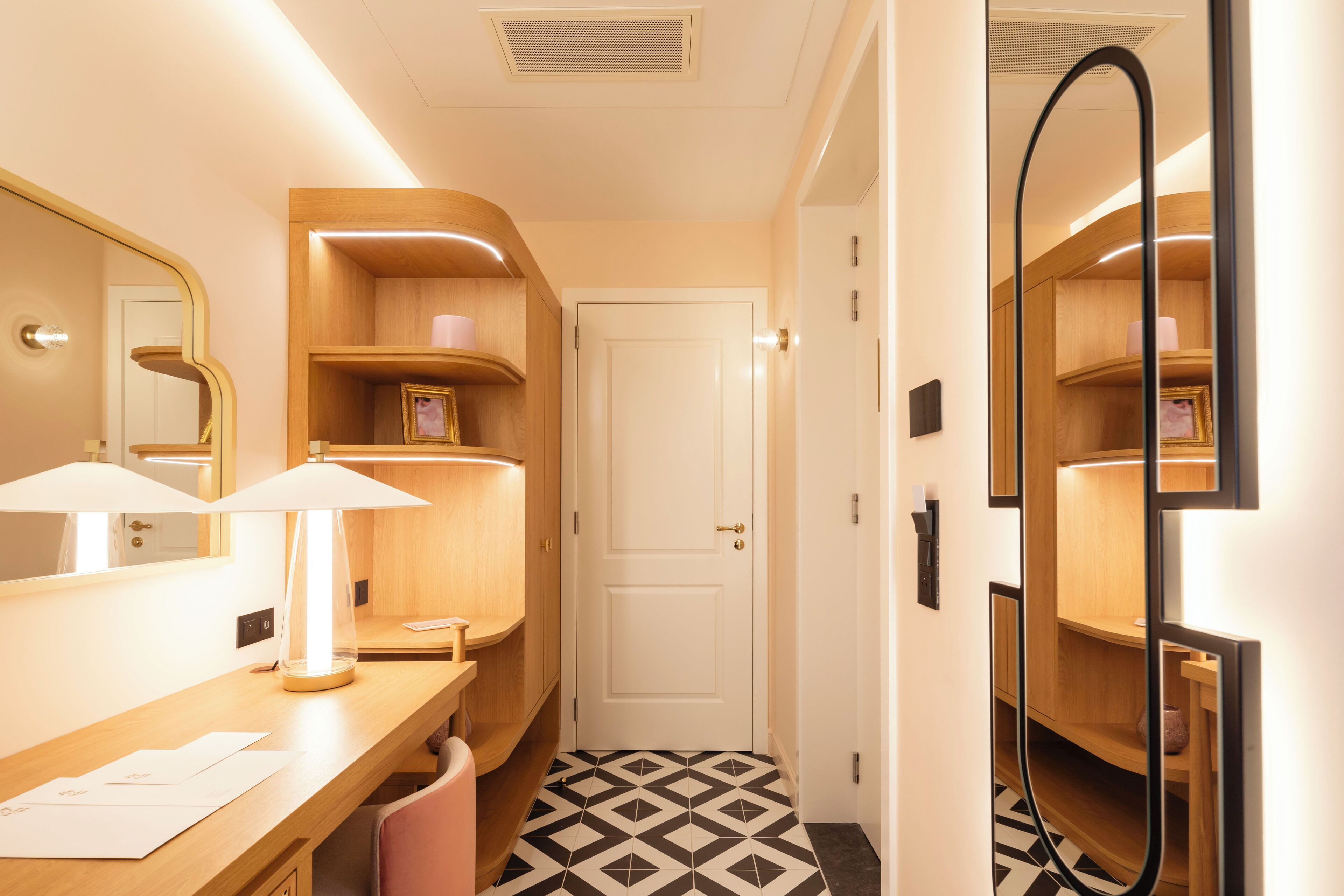 Une chambre d’hôtel avec une coiffeuse en bois, un miroir, un bureau avec une lampe et un sol à motifs.