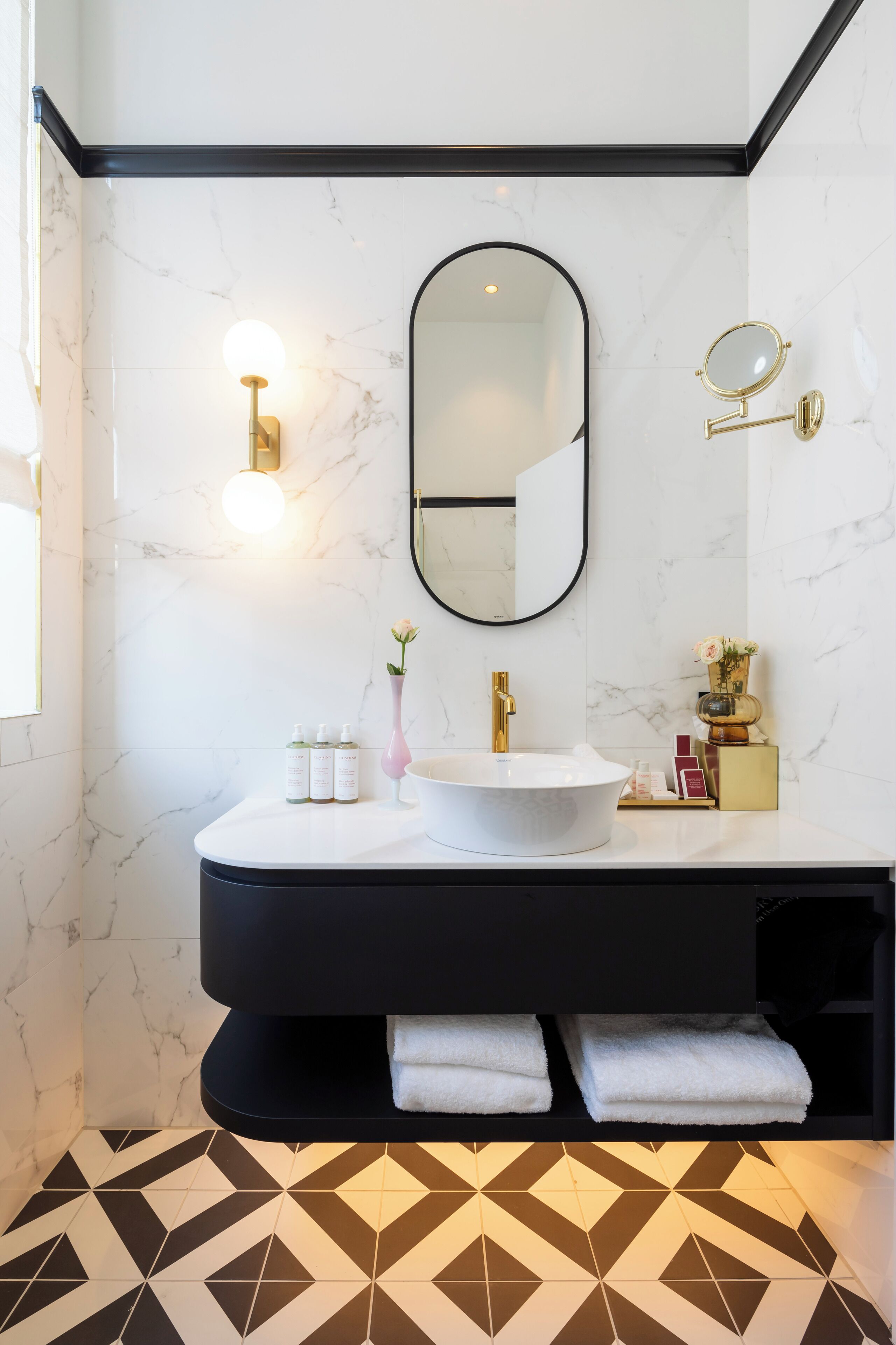 A modern bathroom with a marble wall, a round mirror, a gold faucet, and a geometric patterned floor.
