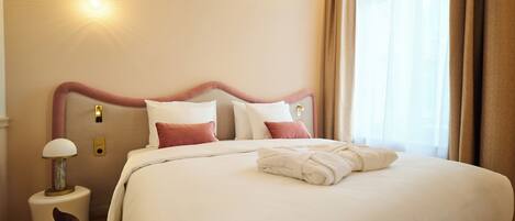 Una cama bien tendida con sábanas blancas y una toalla doblada, una lámpara de noche y una lámpara fijada a la pared.