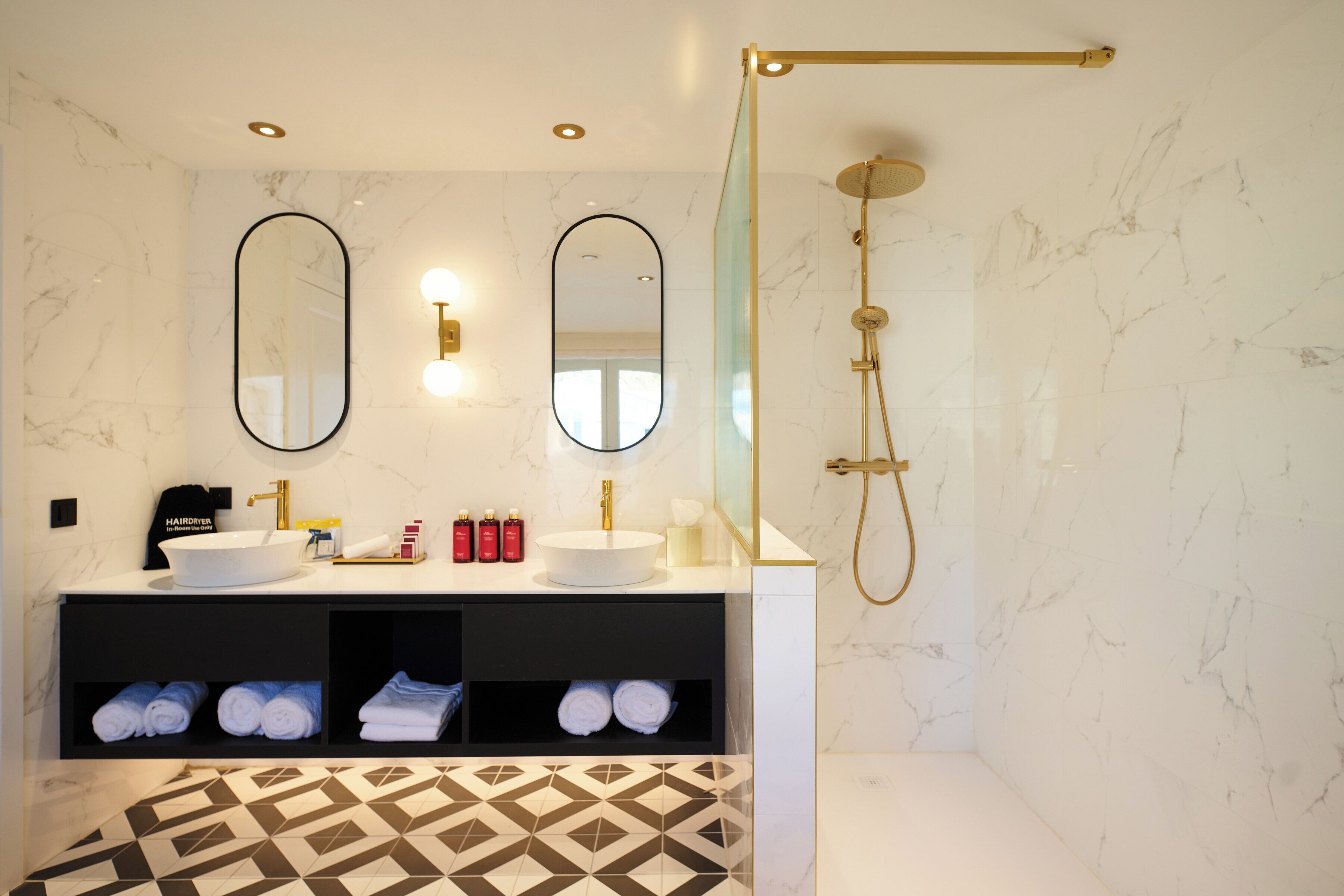 Een moderne badkamer met een grote ovale spiegel, twee wastafels, een inloopdouche en een vloer met een geometrisch patroon.