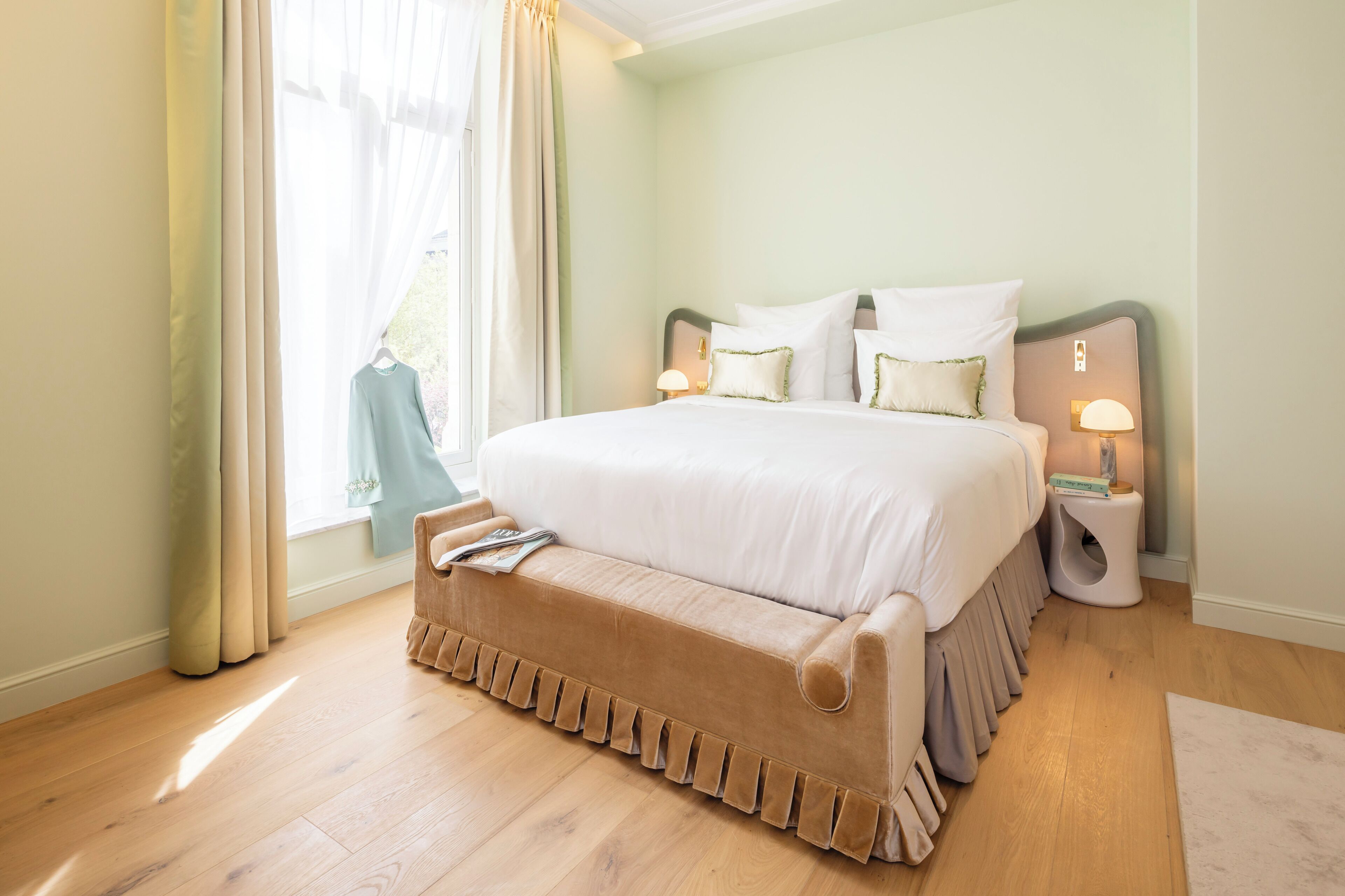 A large bed with a ruffled bed skirt, two bedside lamps, and a window with sheer curtains.