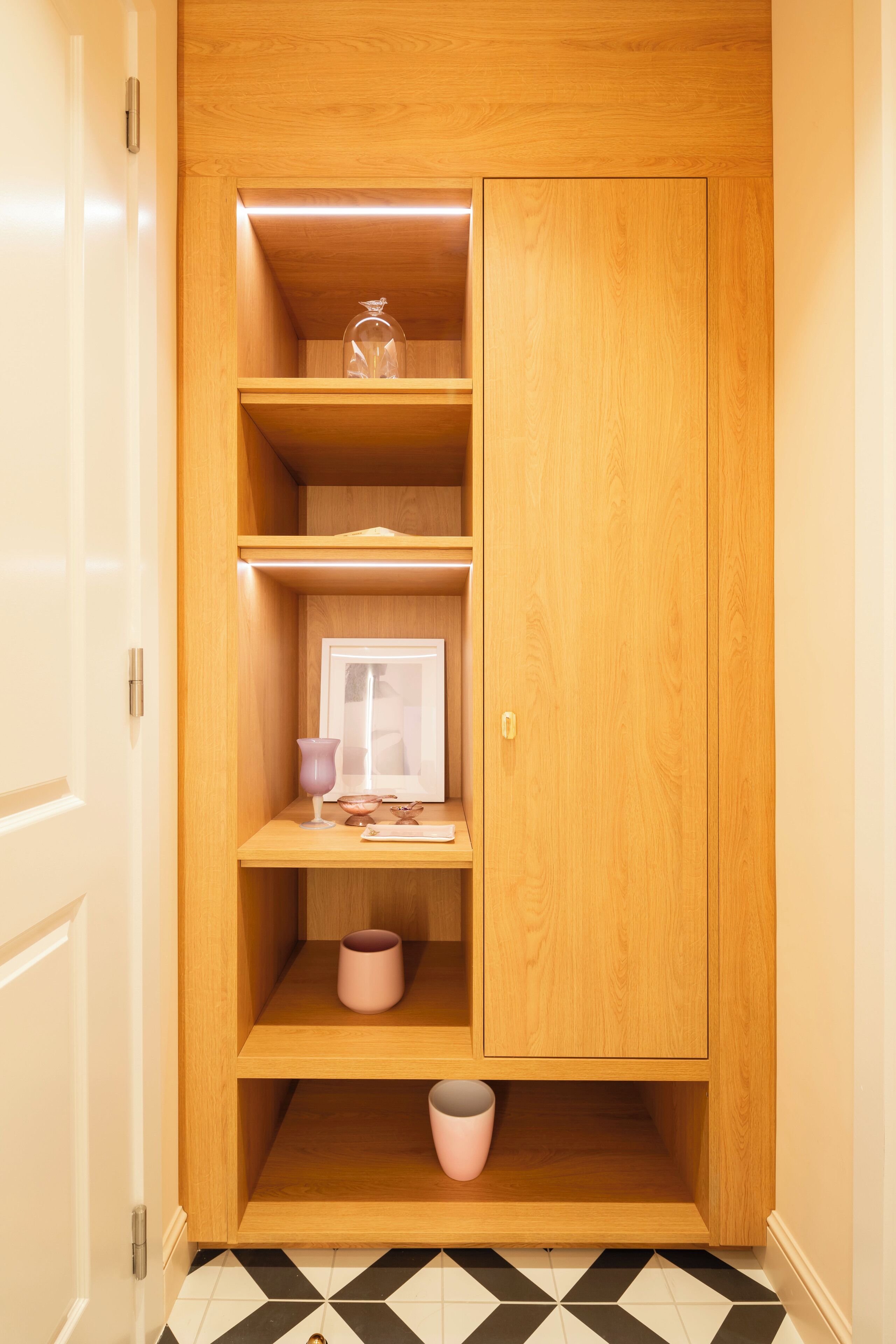 Un armario de madera con varios estantes y una puerta, situado en un pasillo con suelo de baldosas.