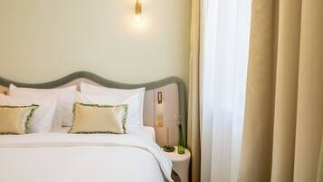 Una cama bien tendida con ropa blanca y almohadas con detalles verdes, una mesita de noche con un jarrón de flores y una lámpara de pared.