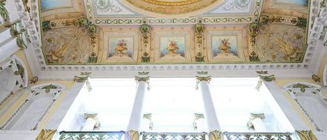 Un salón imponente con frescos ornamentales en el techo, grandes columnas y detalladas decoraciones en las paredes.