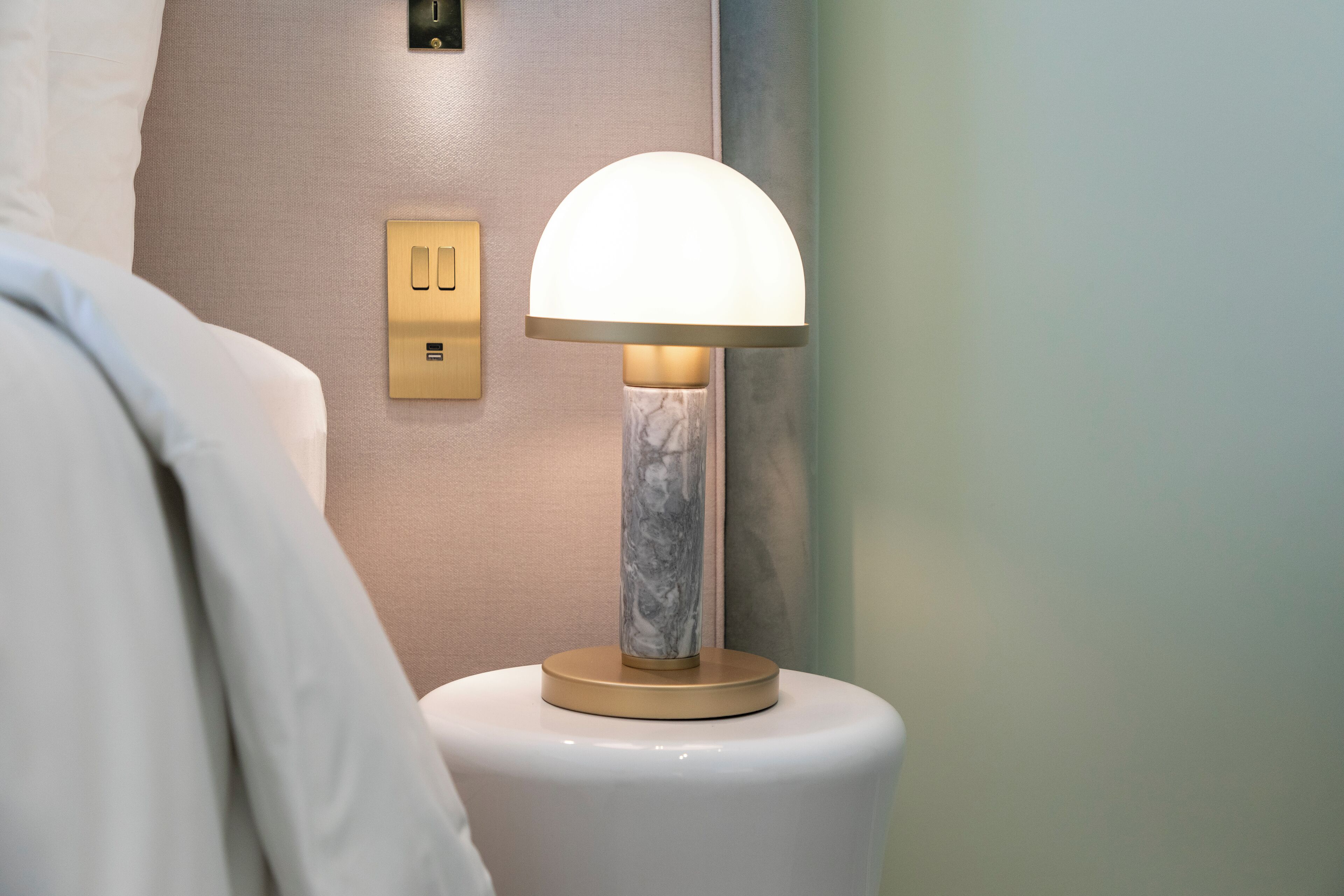 Une table de chevet avec une lampe, une prise électrique murale et un socle qui ressemble à du marbre.