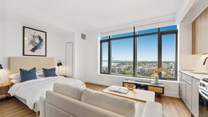 A modern hotel room with a large bed, a sofa, a dining table, and a view of the city through large windows.