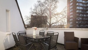Eine Dachterrasse mit rundem Tisch und Stühlen, einer Blumenvase und Blick auf ein Gebäude und Bäume.