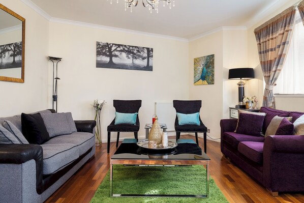 A living room with a grey sofa, a purple sofa, two black armchairs, a glass coffee table, and a green rug. - Luxury Villa 6 Bedrooms Next to Marble Arch (London)