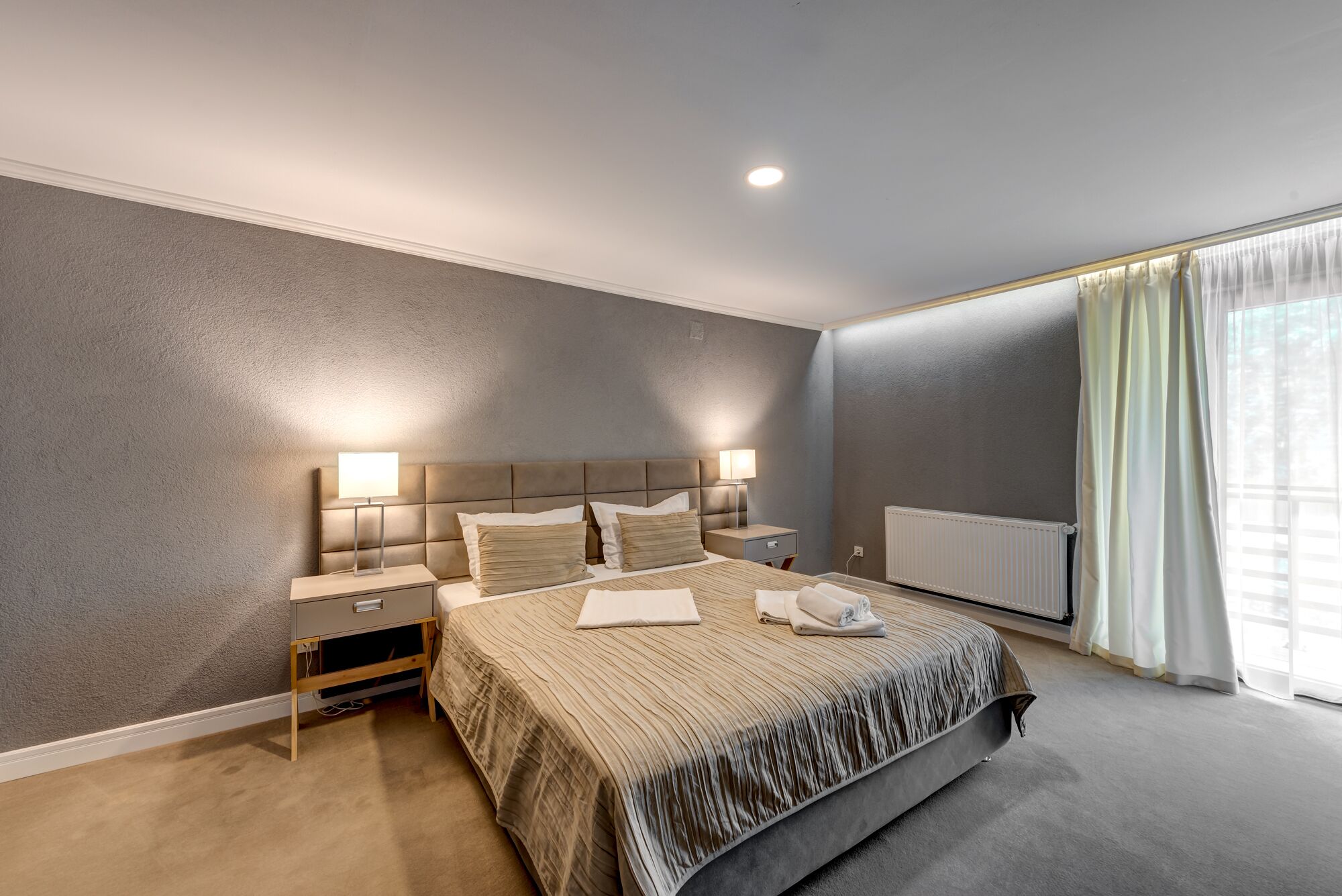Un dormitorio moderno con una cama grande, lámparas de noche, una mesita de noche y una ventana con cortinas.