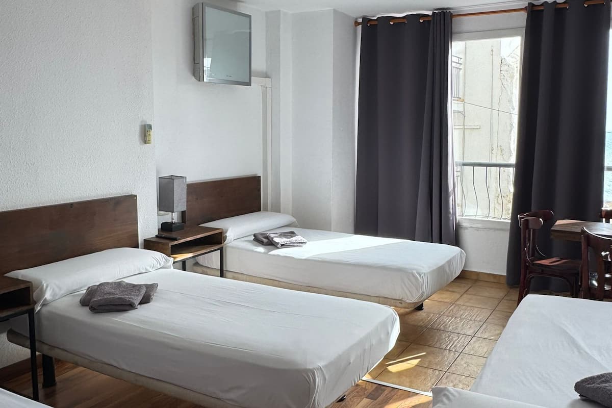 A hotel room with two single beds, a wooden floor, a small desk with a chair, a TV mounted on the wall, and a window with curtains.