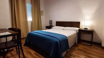 Habitación de hotel con piso de madera, cama con cubrecama azul, escritorio con silla y dos mesitas de noche con lámparas.