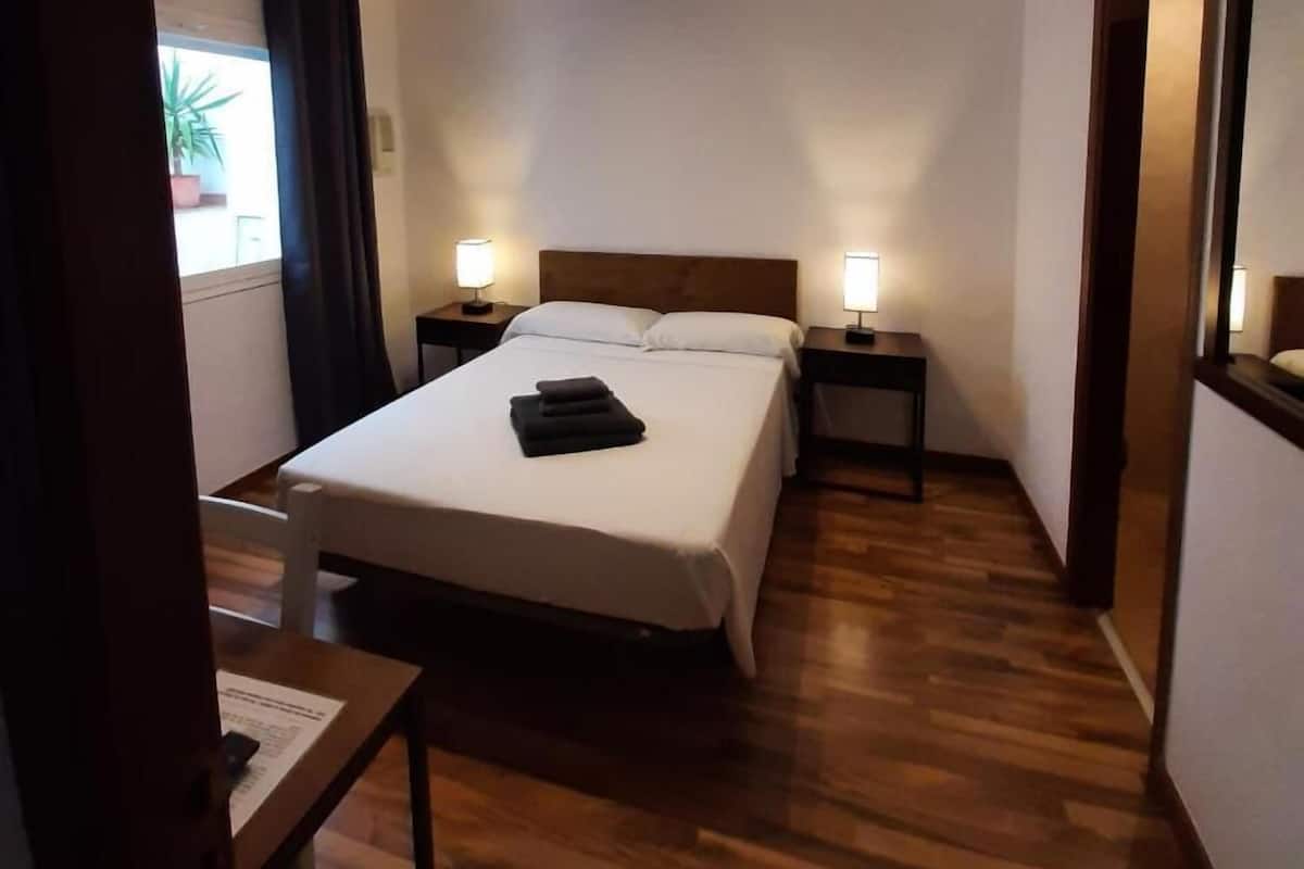 A hotel room with a bed, two bedside tables with lamps, a wooden floor, and a window with curtains.