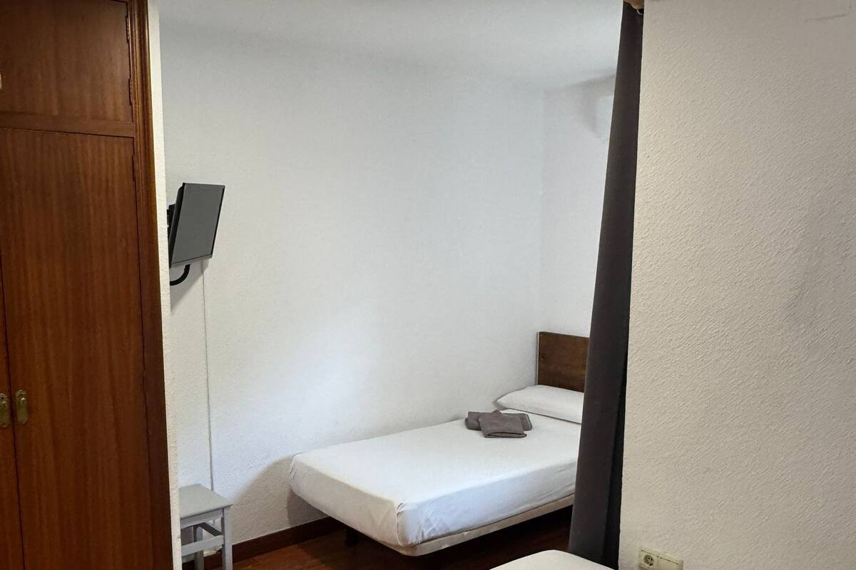A small, single-room accommodation with a wooden floor, a single bed, a small table, and a wall-mounted TV.