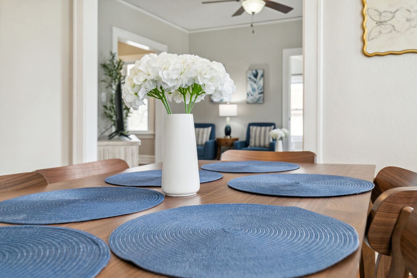 A dining table with blue placemats and a white vase with white flowers.