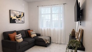 Ein Wohnzimmer mit einem dunkelgrauen Ecksofa, einem Boden aus weißem Marmorfliesen, einem großen Fenster mit transparenten Vorhängen, einem fest montierten Flachbildfernseher und einem Wandregal mit Dekoration.