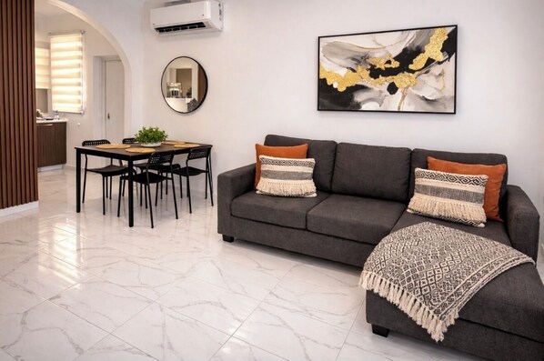 Una sala de estar moderna con un sofá gris, dos almohadones estampados, un espejo redondo y un cuadro abstracto enmarcado.