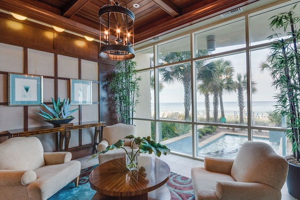 A spacious room with a large window overlooking a pool and palm trees, featuring a wooden ceiling, a round wooden table, and several chairs.