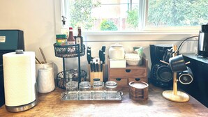 En køkkenbordplade med forskellige ting, heriblandt et køkkenrulleholder, krukker, en kaffemaskine og et vindue med udsigt til grøn natur.