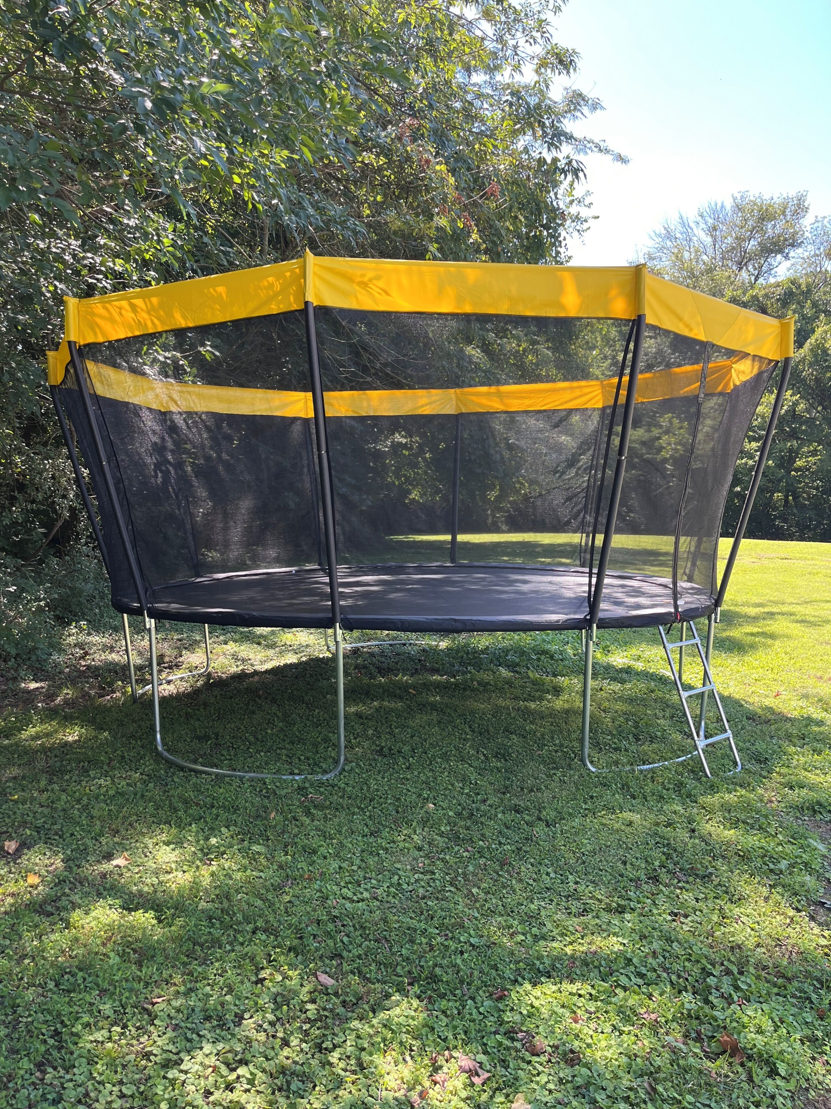 Un trampolín con red de seguridad amarilla, rodeado de césped y árboles.