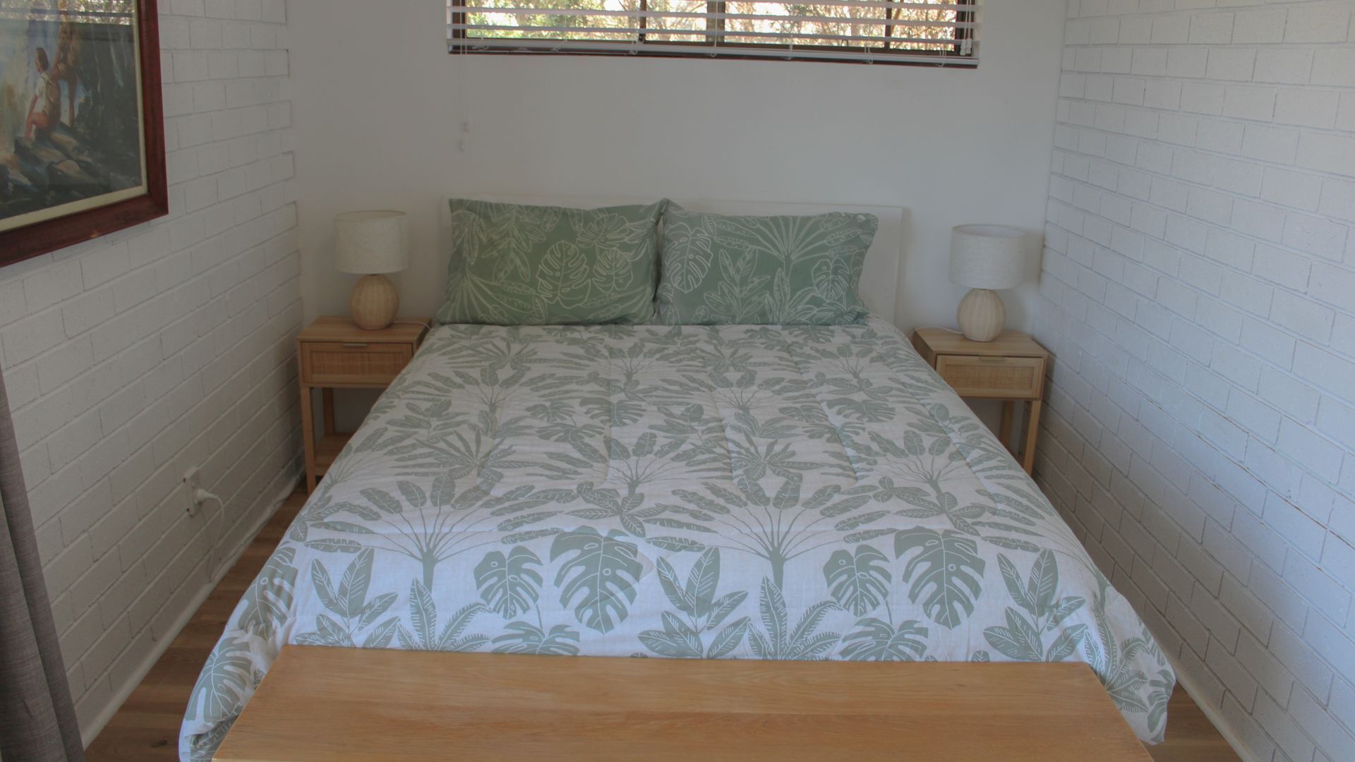 A bedroom with a bed, two nightstands, two lamps, a window with blinds, and a painting on the wall.