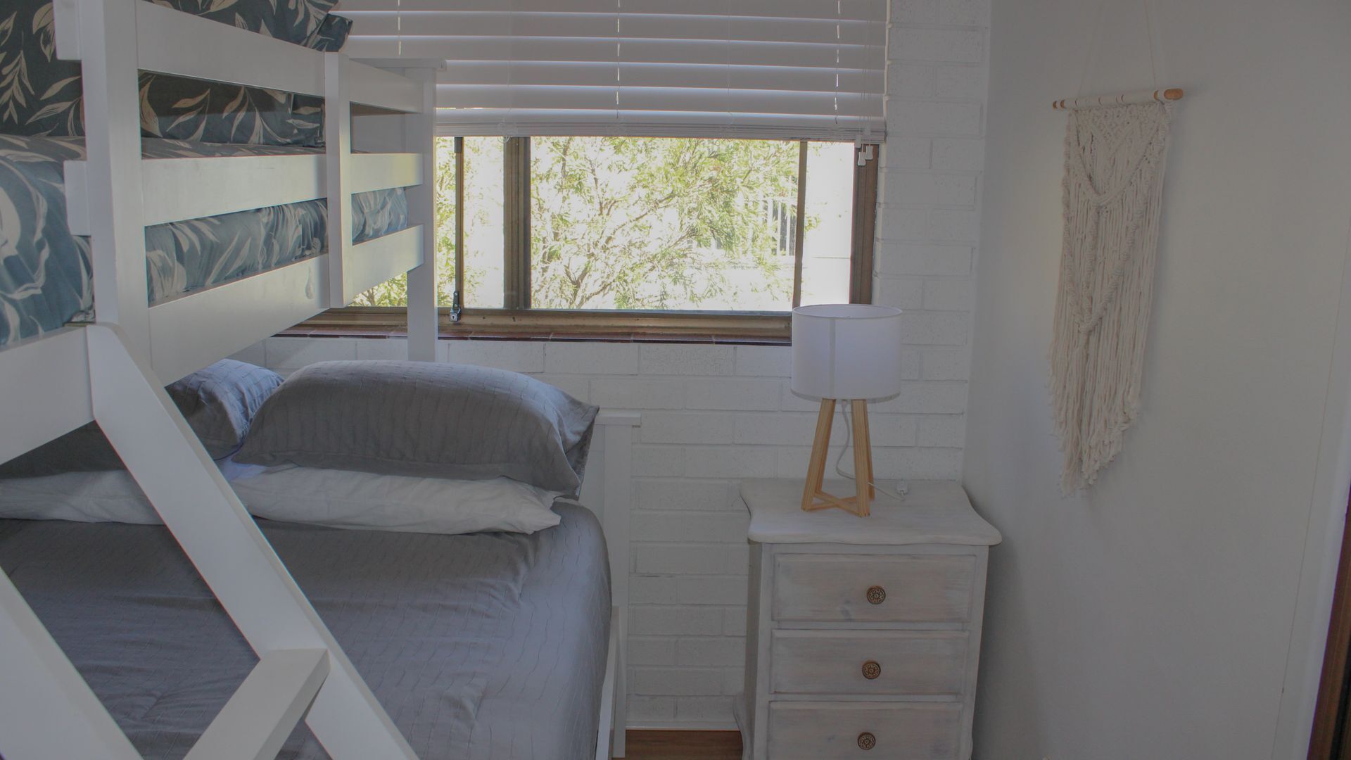 A bunk bed with a window, a bedside table with a lamp, and a white wall with a textured curtain.