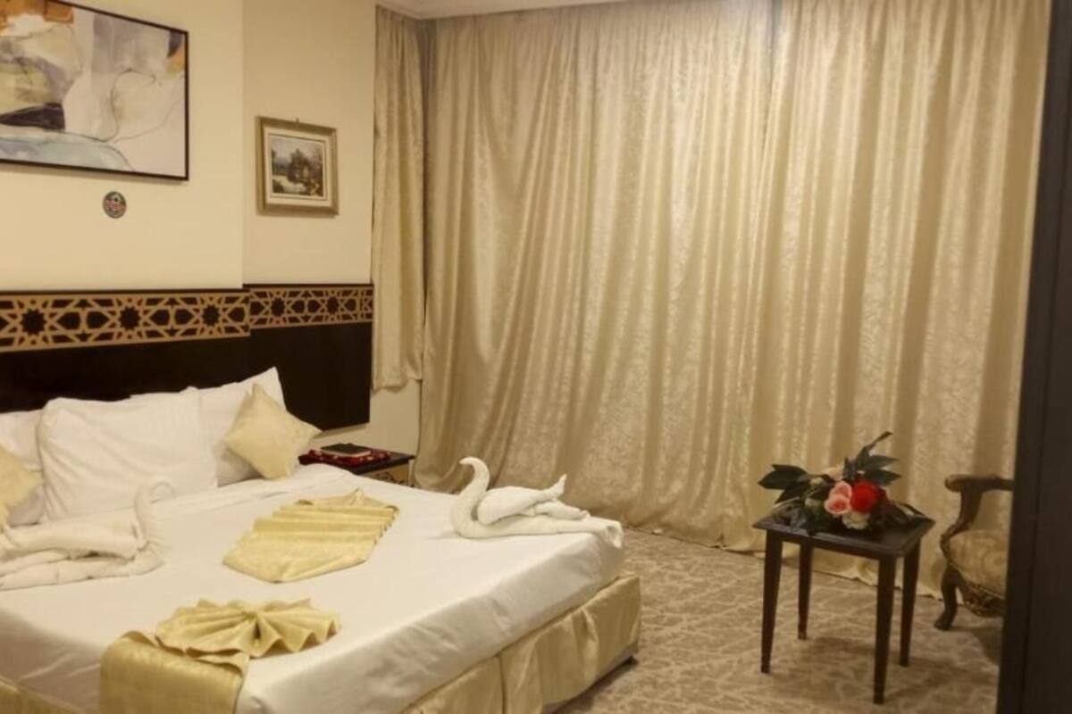 Ein Hotelzimmer mit einem ordentlich bezogenen Bett, einem kleinen Tisch mit Blumen in einer Vase und einem großen Fenster mit Vorhängen.