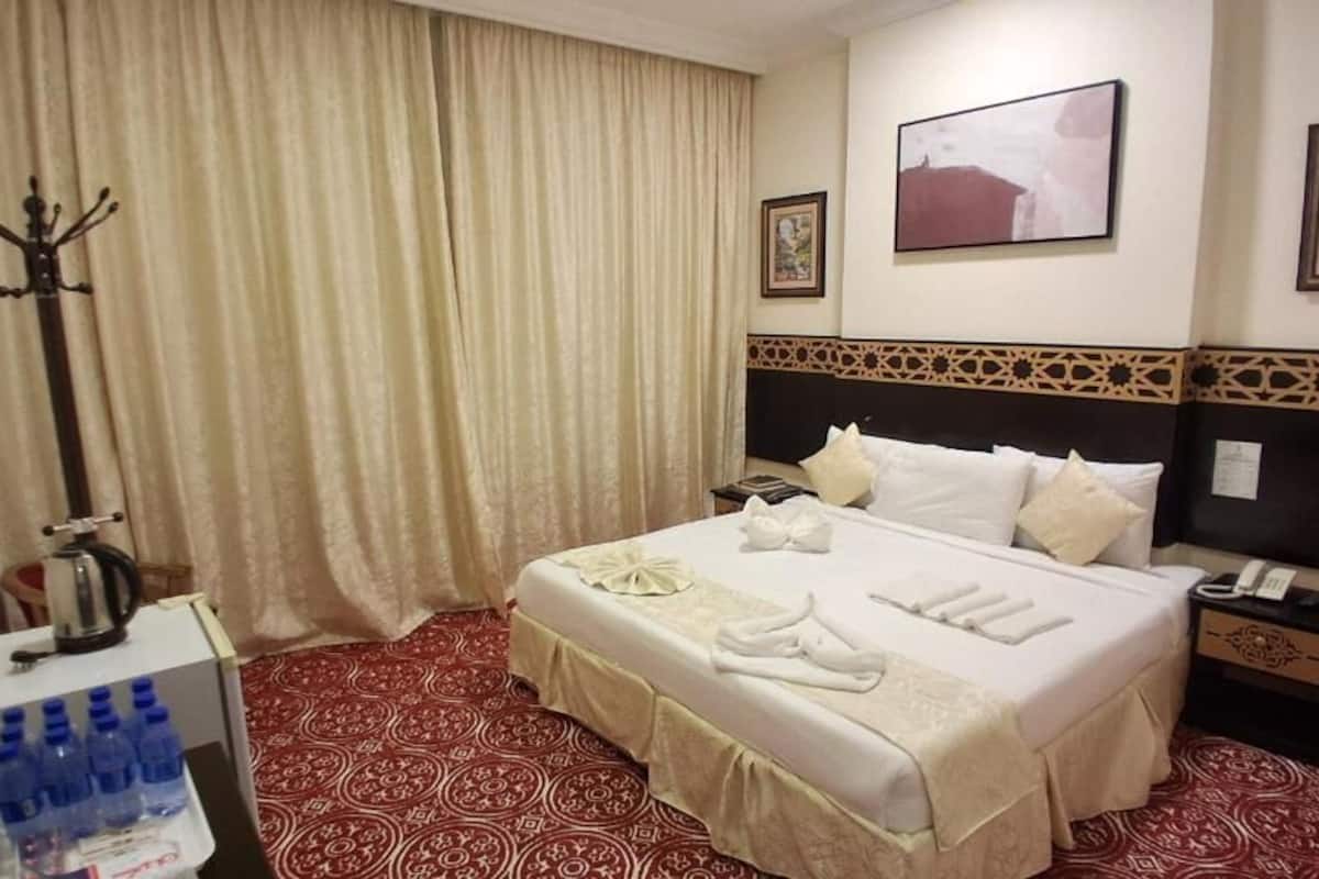 Ein Hotelzimmer mit einem großen Bett, einem Nachttisch mit Lampe, einem an der Wand befestigten Fernseher und einem Fenster mit Vorhängen.